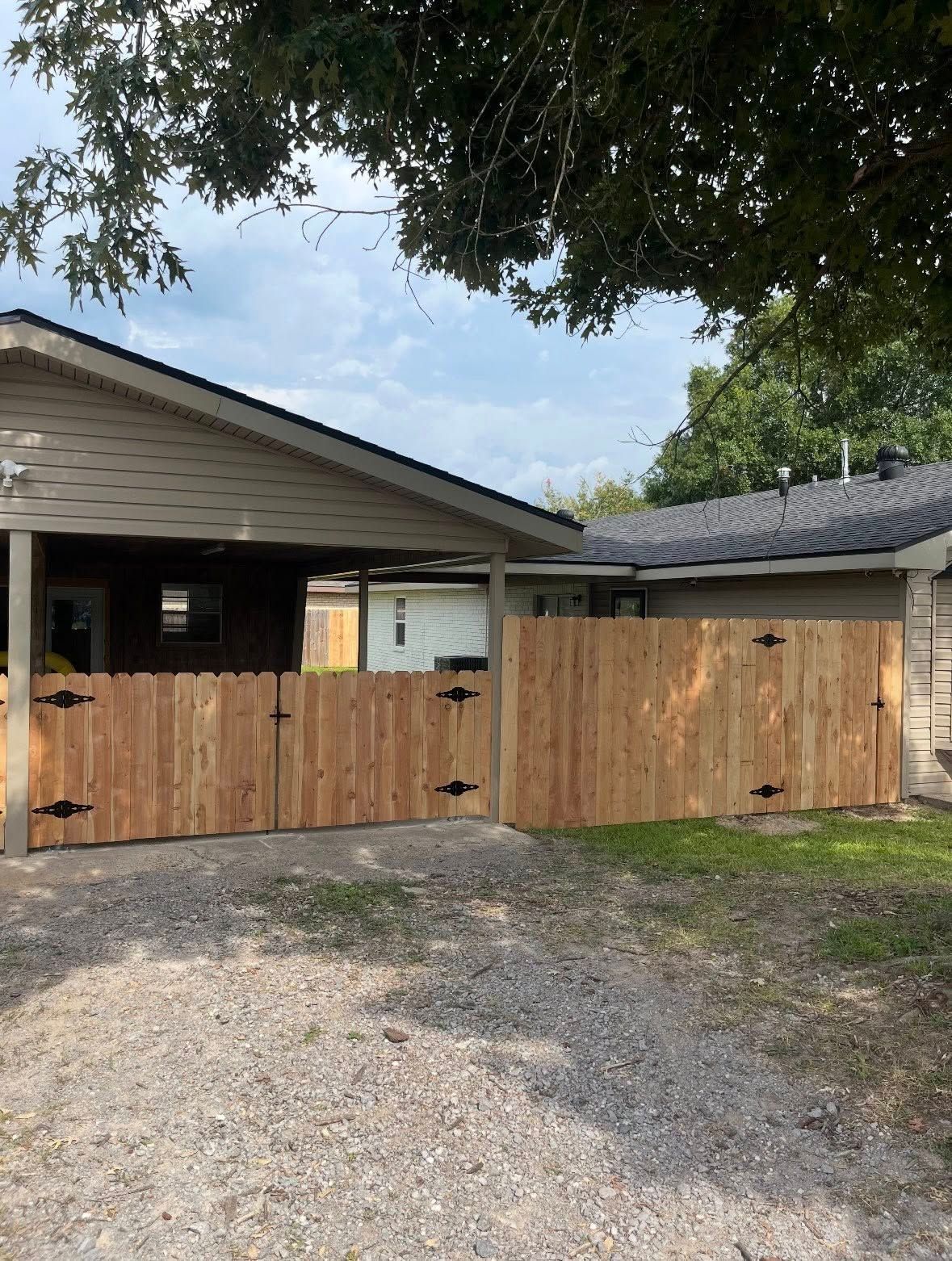 Wooden driveway gate beside a house, with a gravel drive and trees overhead.