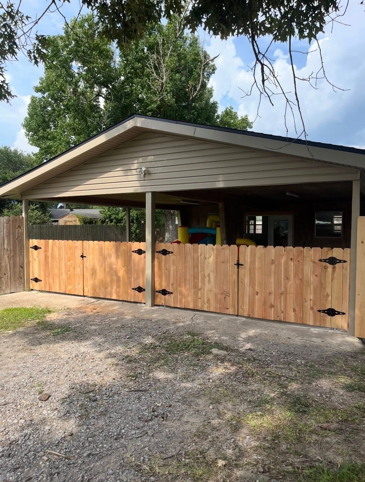 Wooden fence and carport on a gravel driveway beneath trees on a sunny day