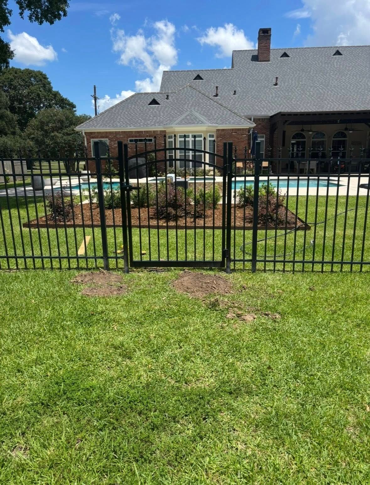 Black metal fence around a landscaped yard with a house and cloudy blue sky in the background