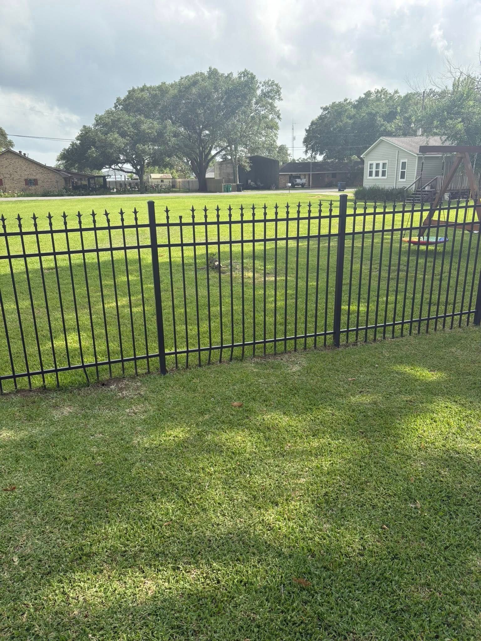 Black metal fence around a grassy yard with trees and houses in the background under a cloudy sky