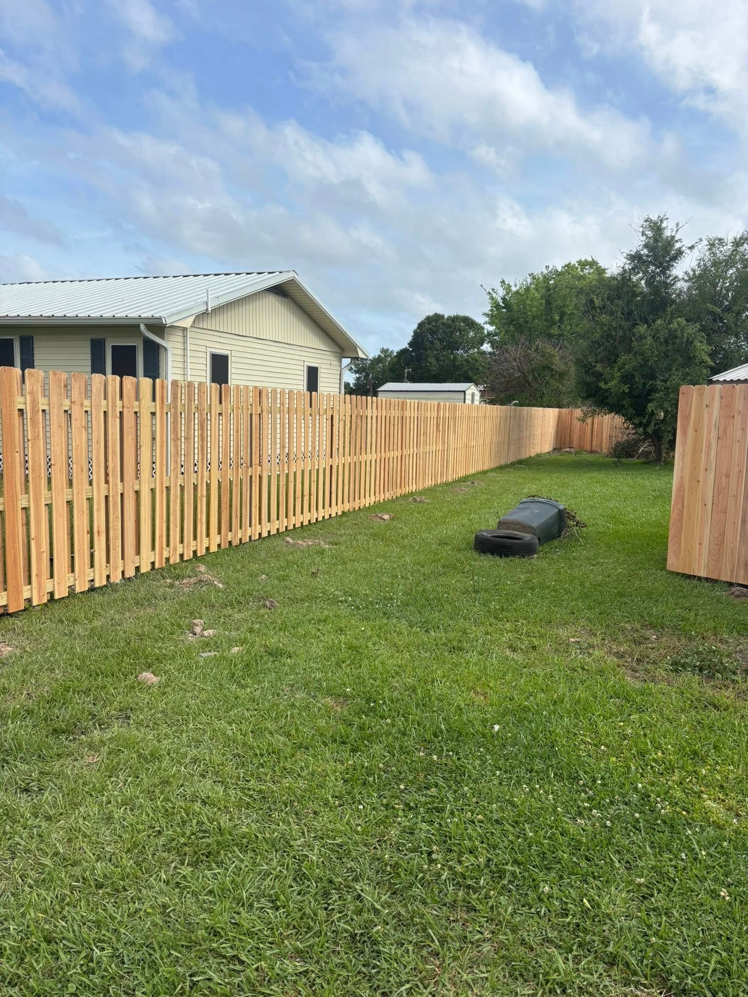 Backyard lawn with a wooden fence, house, and a black grill cover on the grass under a cloudy sky
