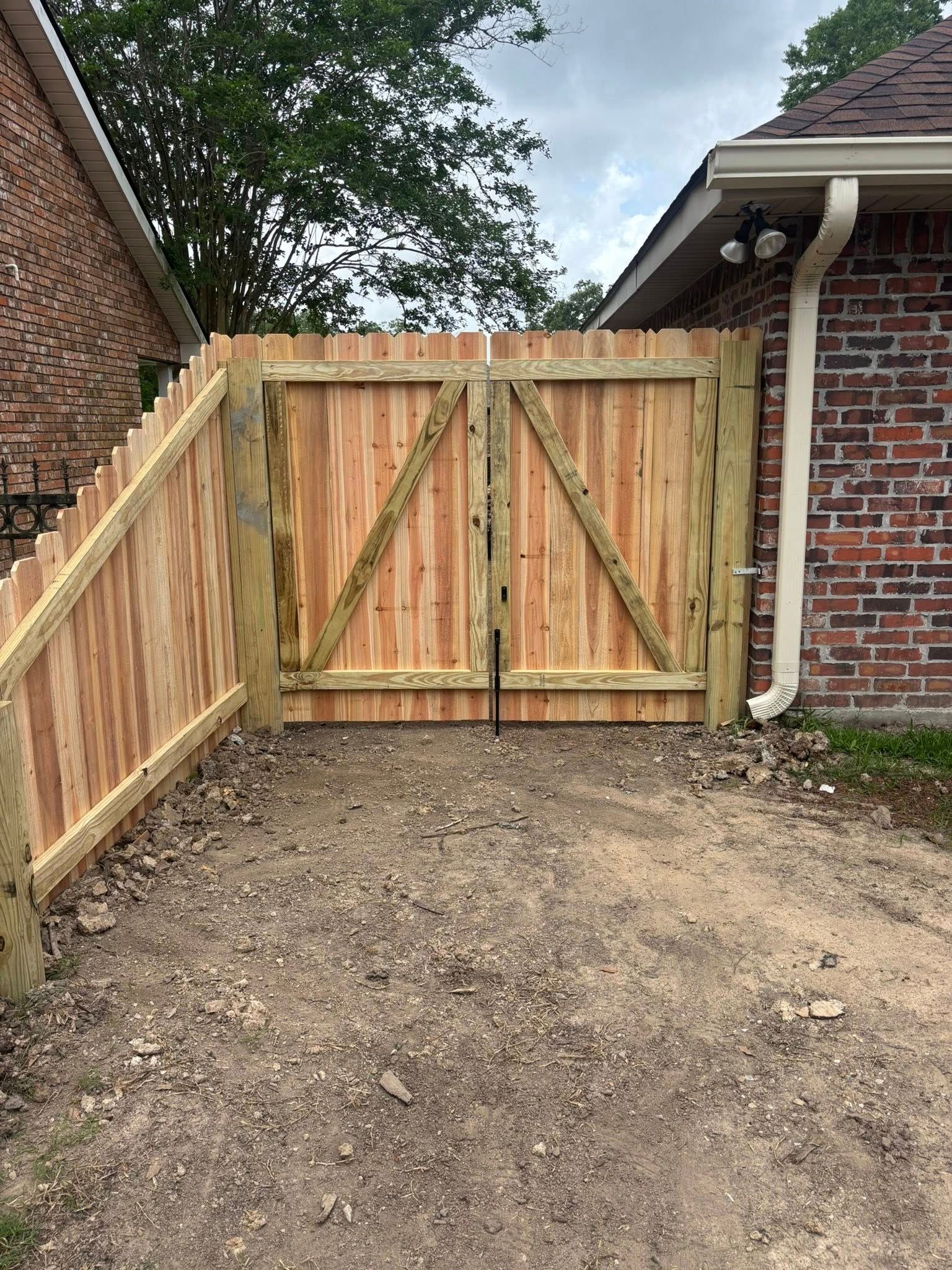 Wooden driveway gate with diagonal brace, between brick walls and a dirt yard.