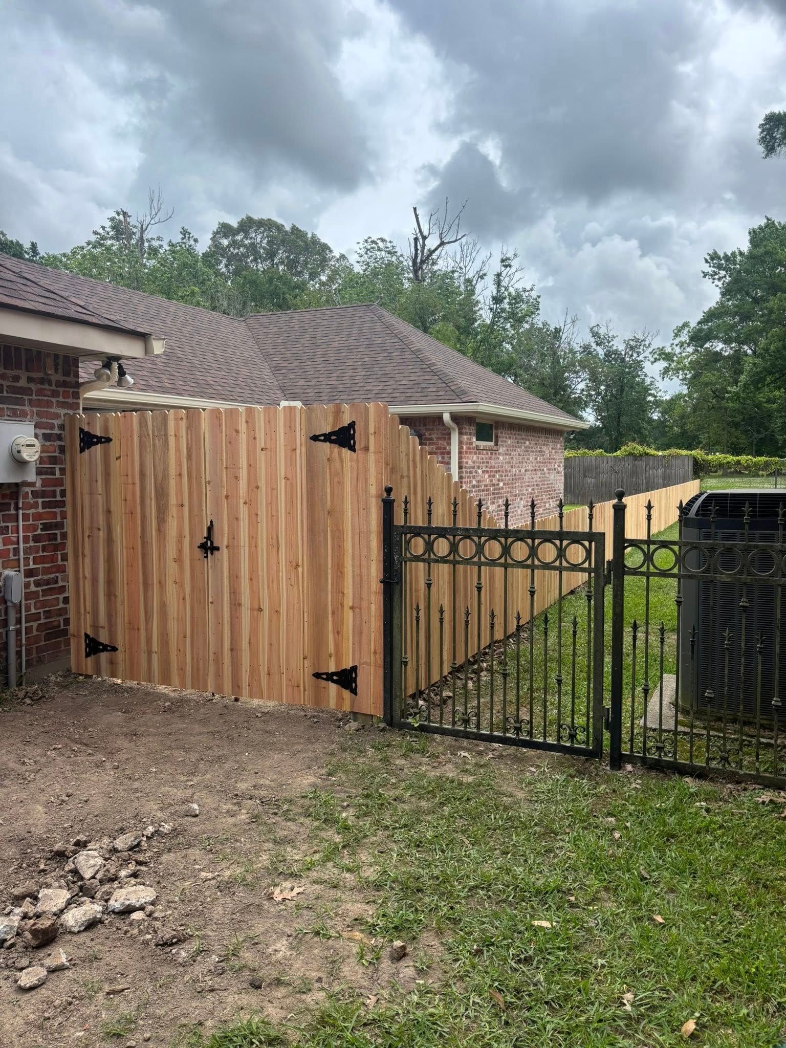 Wooden gate and black metal fence beside a house under cloudy skies, with muddy ground and grass.