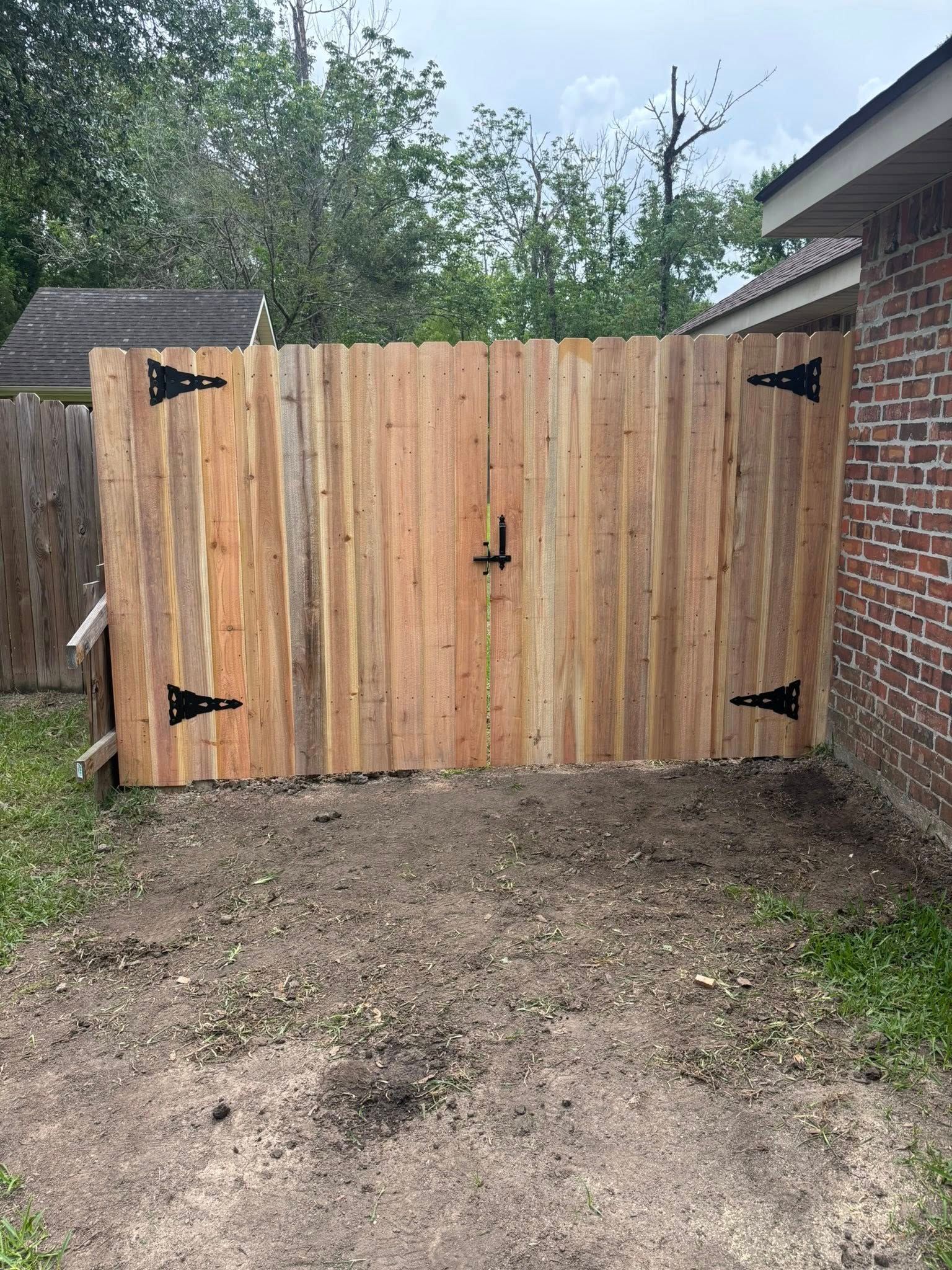 Wooden double gate with black hinges in a brick-and-fence backyard entrance