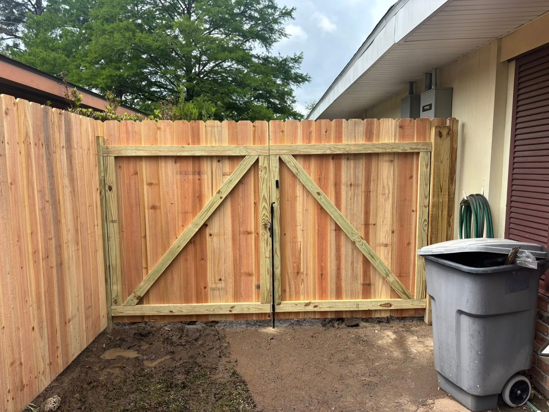 Wooden backyard gate between fences beside a house, with a trash bin on the right.