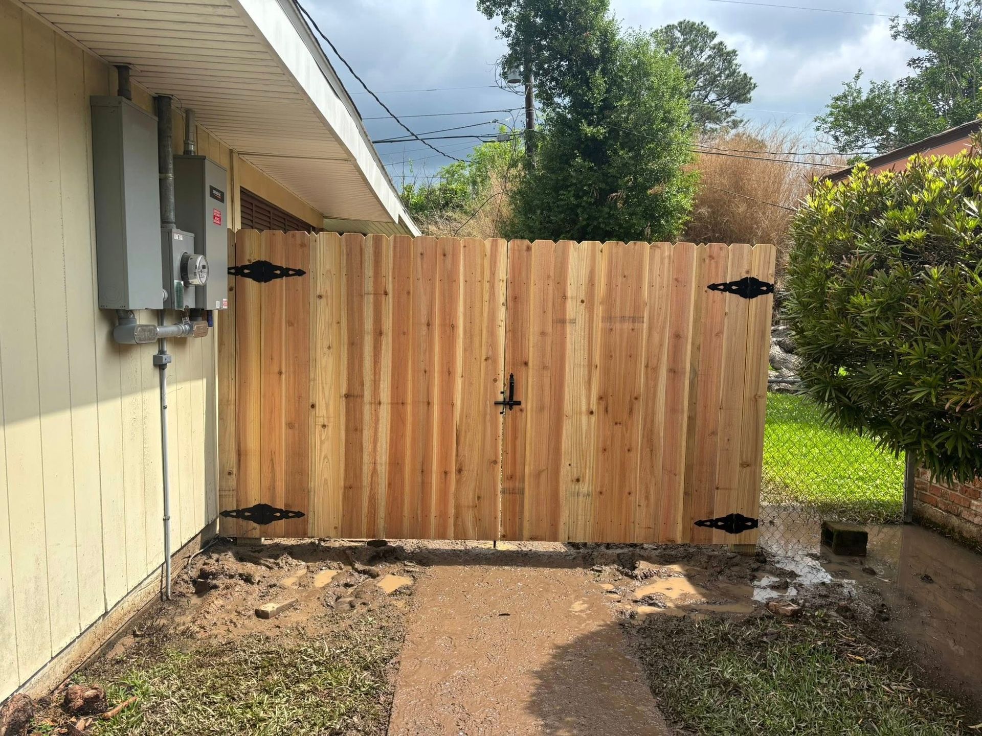 Wooden gate blocking a narrow side yard beside a beige house, with greenery and gravel path.