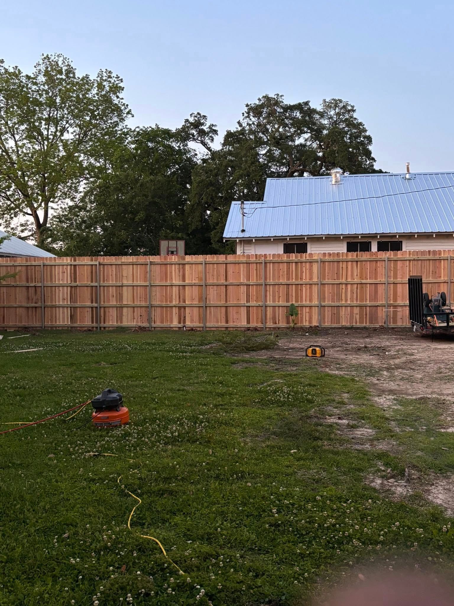Backyard with a wooden privacy fence, grassy yard, trees, and a partially roofed house in the background