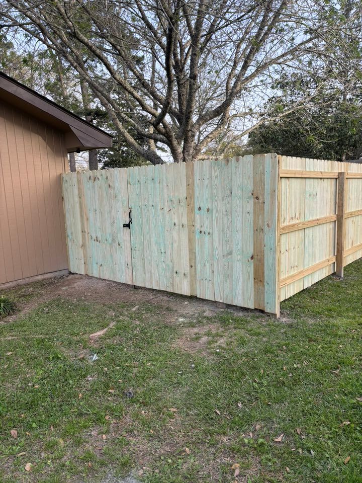 Weathered wooden fence panels beside a beige building, with a large tree and grassy yard behind it.