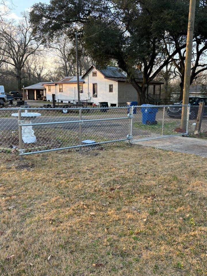 Chain-link fenced yard with a small white house, trees, and a blue trash bin.