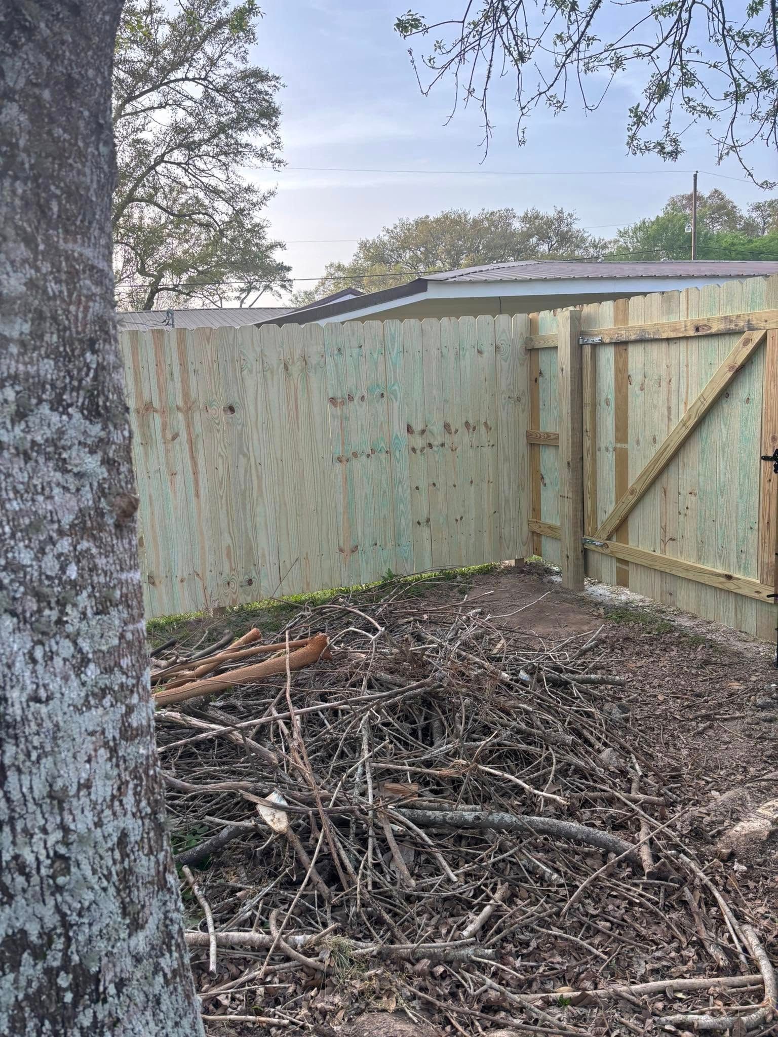 Backyard corner with wooden fence, gate, tree trunk, and a pile of branches and yard debris on the ground