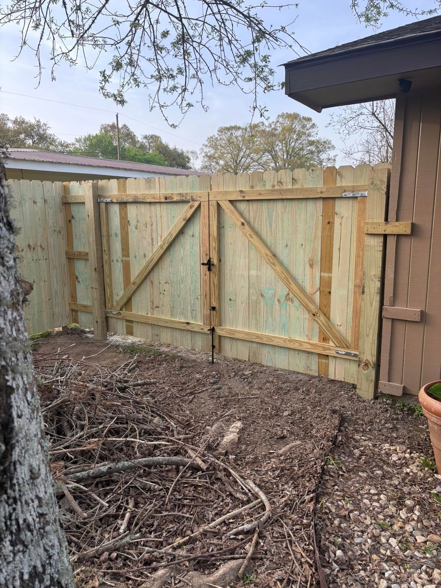 Wooden backyard gate with diagonal braces, leaves and branches on the ground, beside a house and fence.