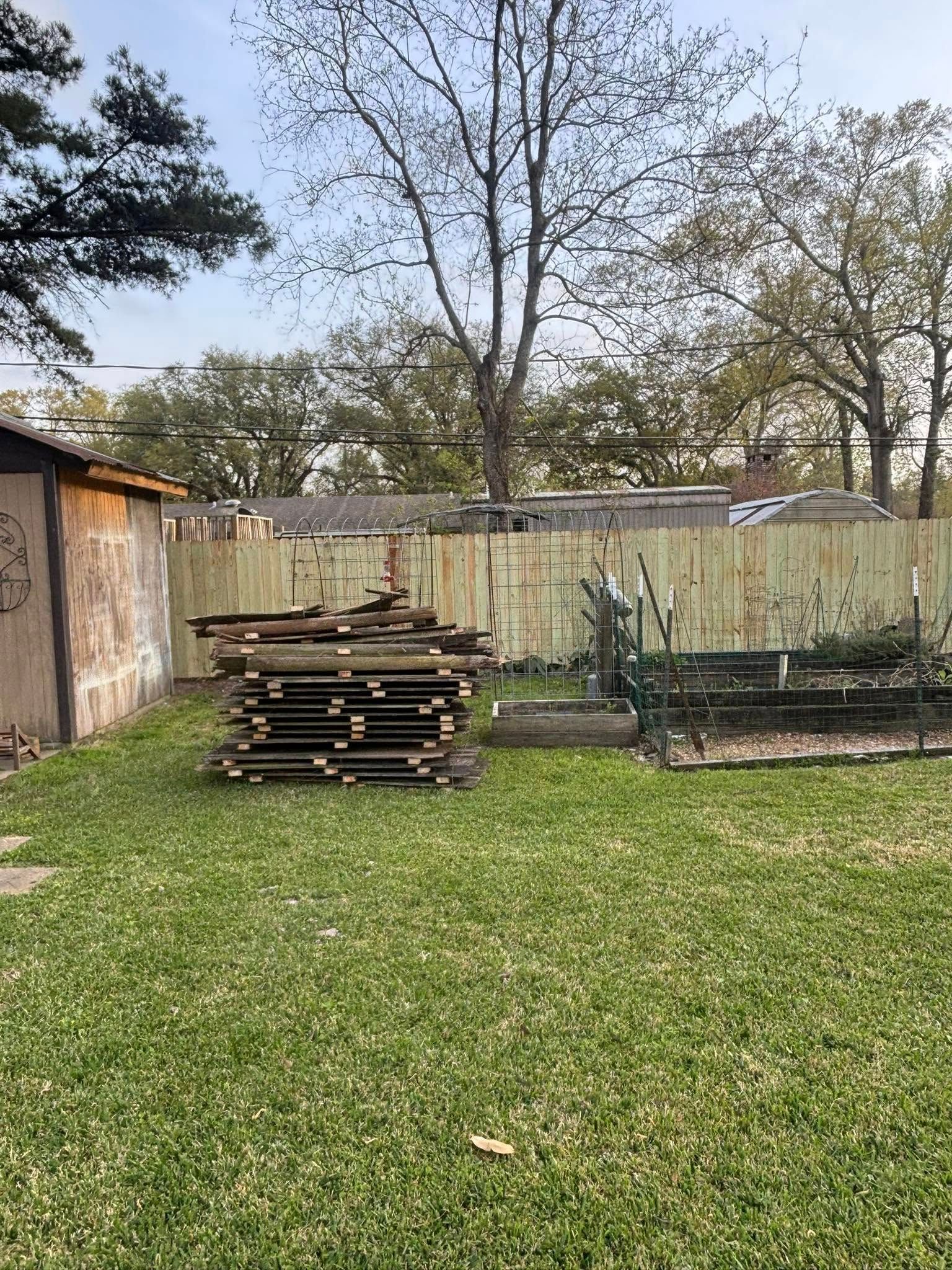 Backyard with wooden fence, stacked boards, and a small garden bed on grass.