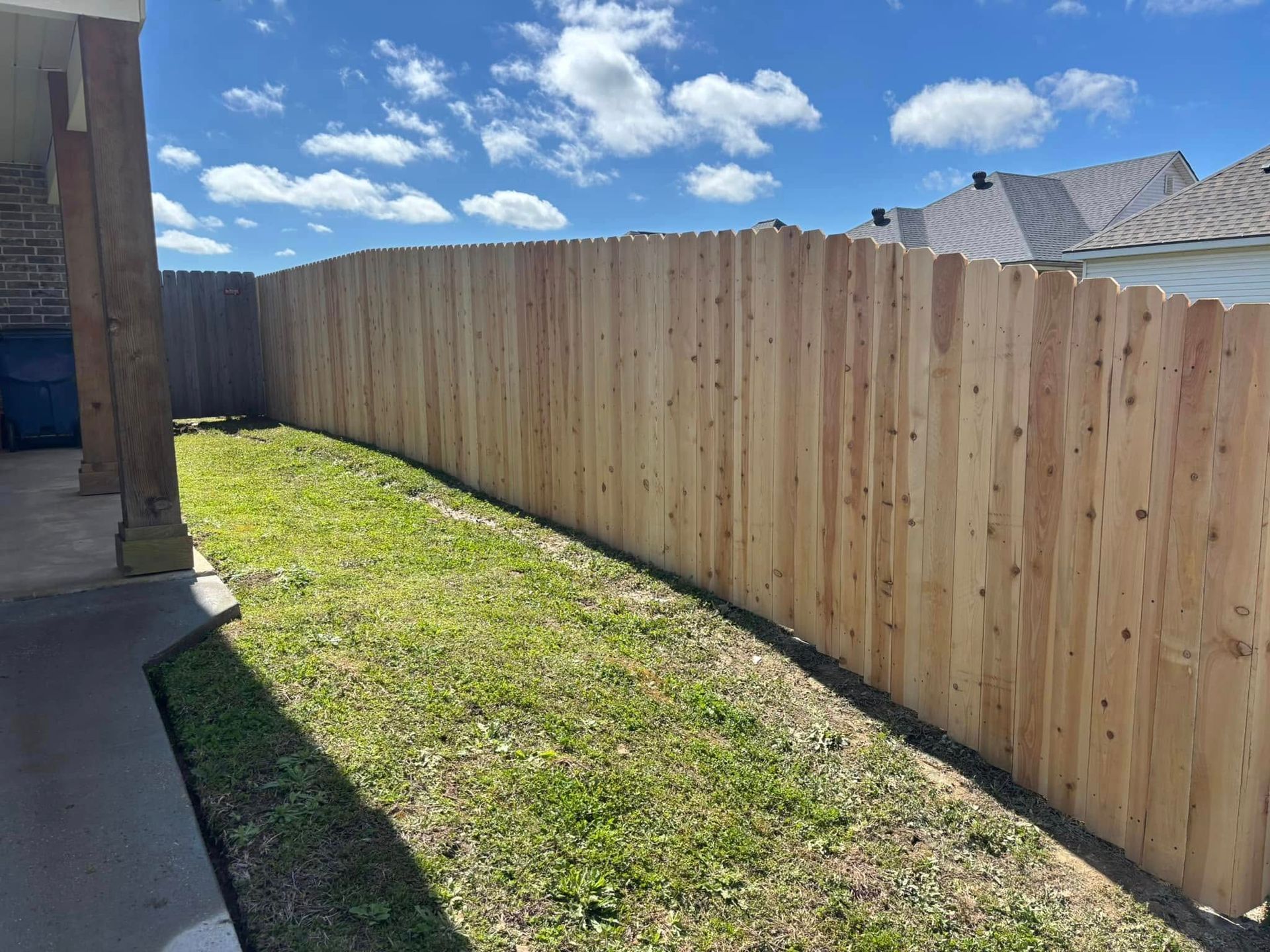 Narrow grassy side yard beside a house, bordered by a tall wooden fence under a blue sky.