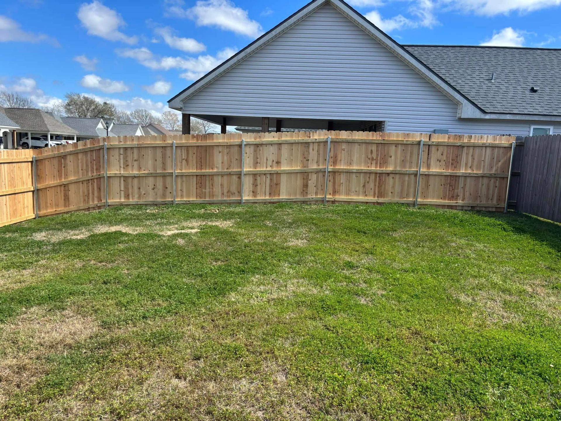 Backyard with a wooden fence, patchy green grass, and a gray house under a blue sky.
