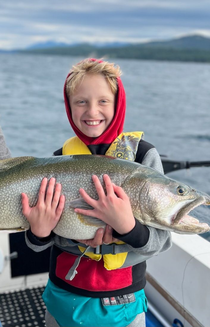 Boy smiling, holding a large fish on a boat, lake and mountains in the background.