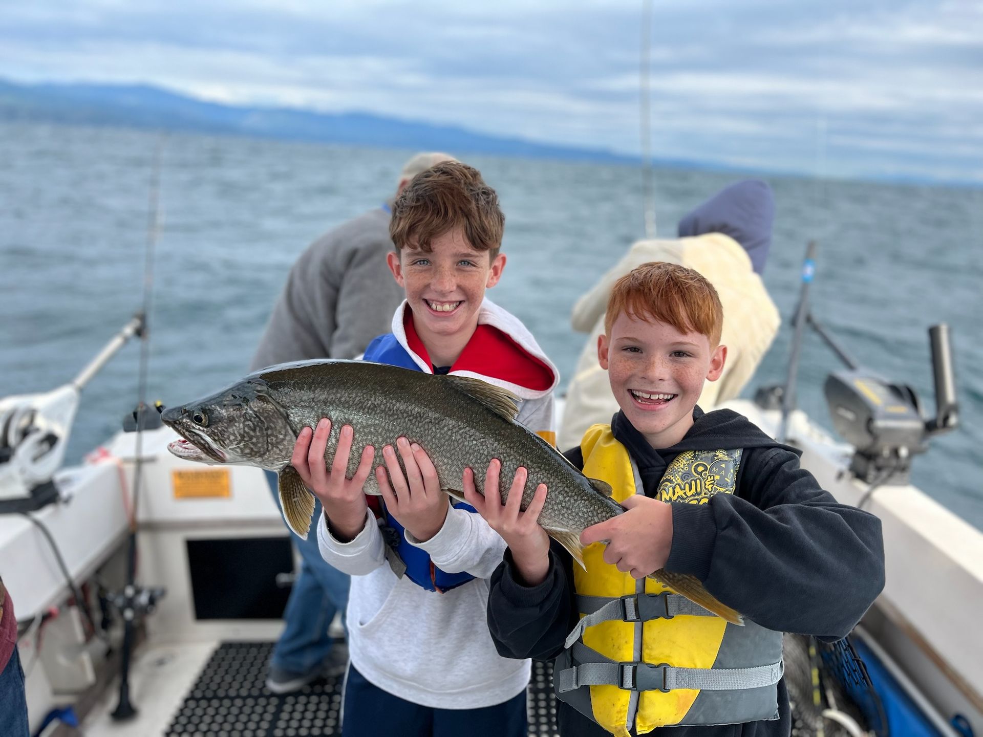 Two boys on a boat hold a large fish. They smile, wearing life vests, with water and mountains in the background.