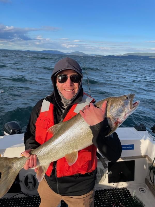 Man on a boat holding a large fish, smiling. Overcast sky, water in the background.