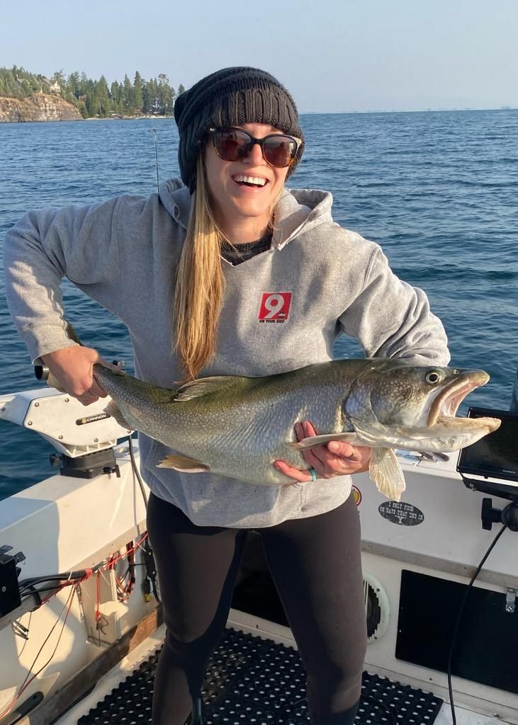Woman on a boat holding a large fish, smiling. She wears a beanie, sunglasses, and a gray hoodie. Lake in the background.