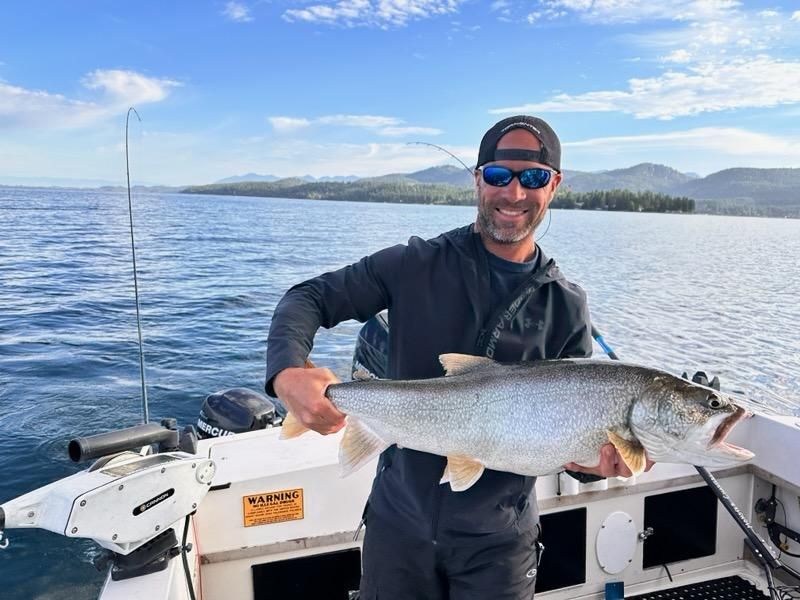 Man on a boat holds up a large, silver fish he caught, smiling, with lake and sky backdrop.