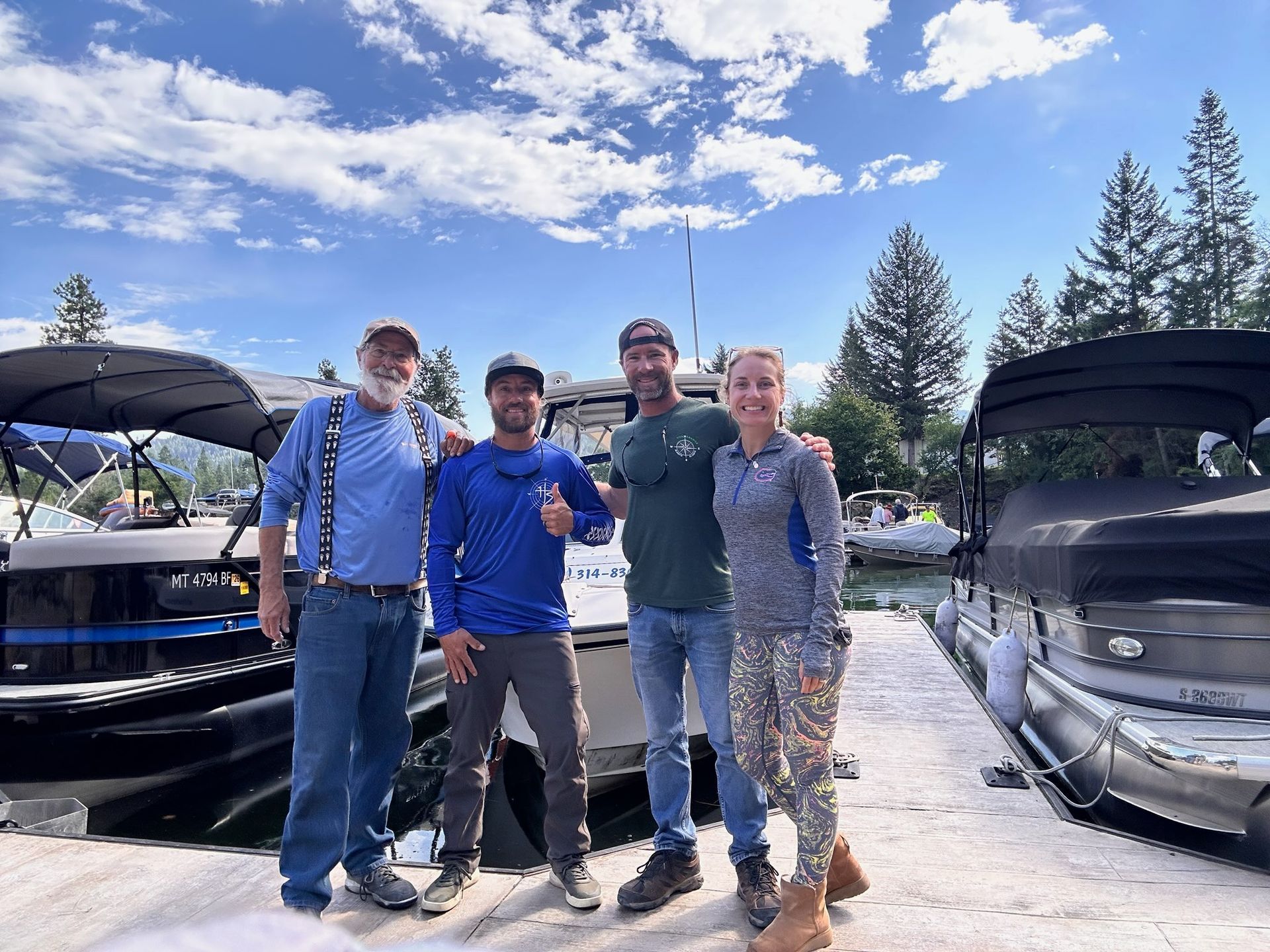 Four people stand by boats on a sunny day. Two have arms around each other, smiling.