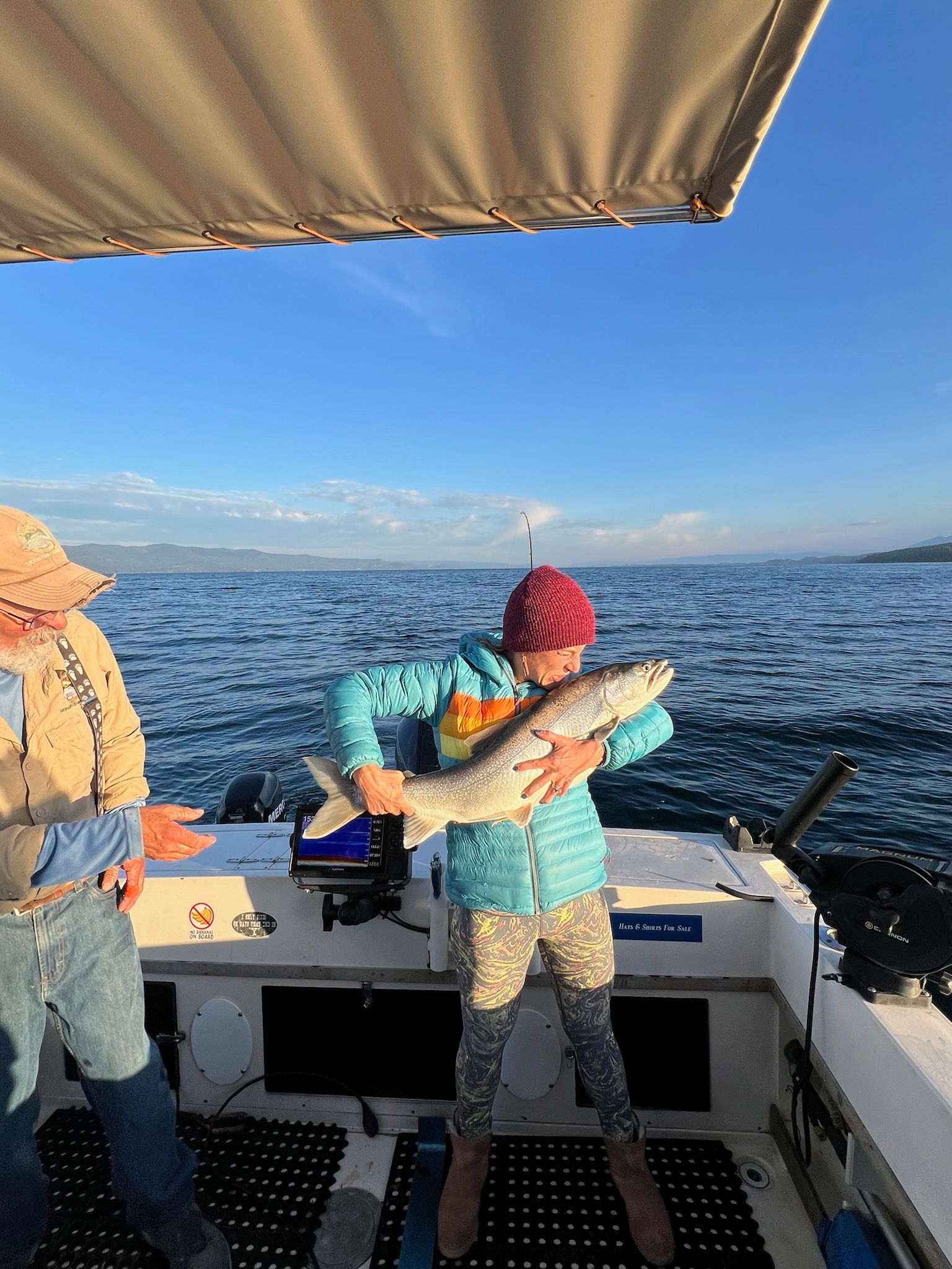 Person on a boat holding a fish, with another person nearby. Blue water and sky in the background.
