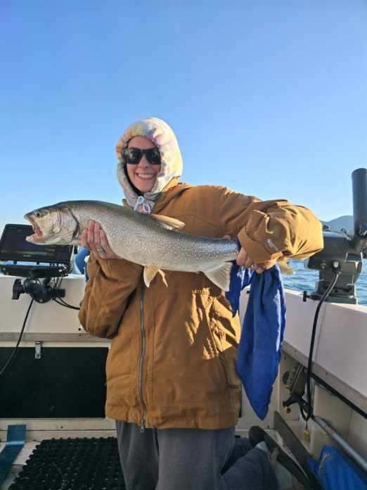 Woman in a hooded jacket smiling, holding a large fish on a boat.