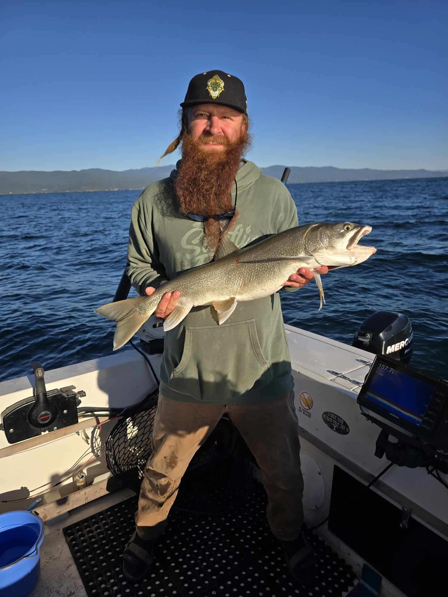Man on boat holds up a large fish he caught, clear water, mountains in background.