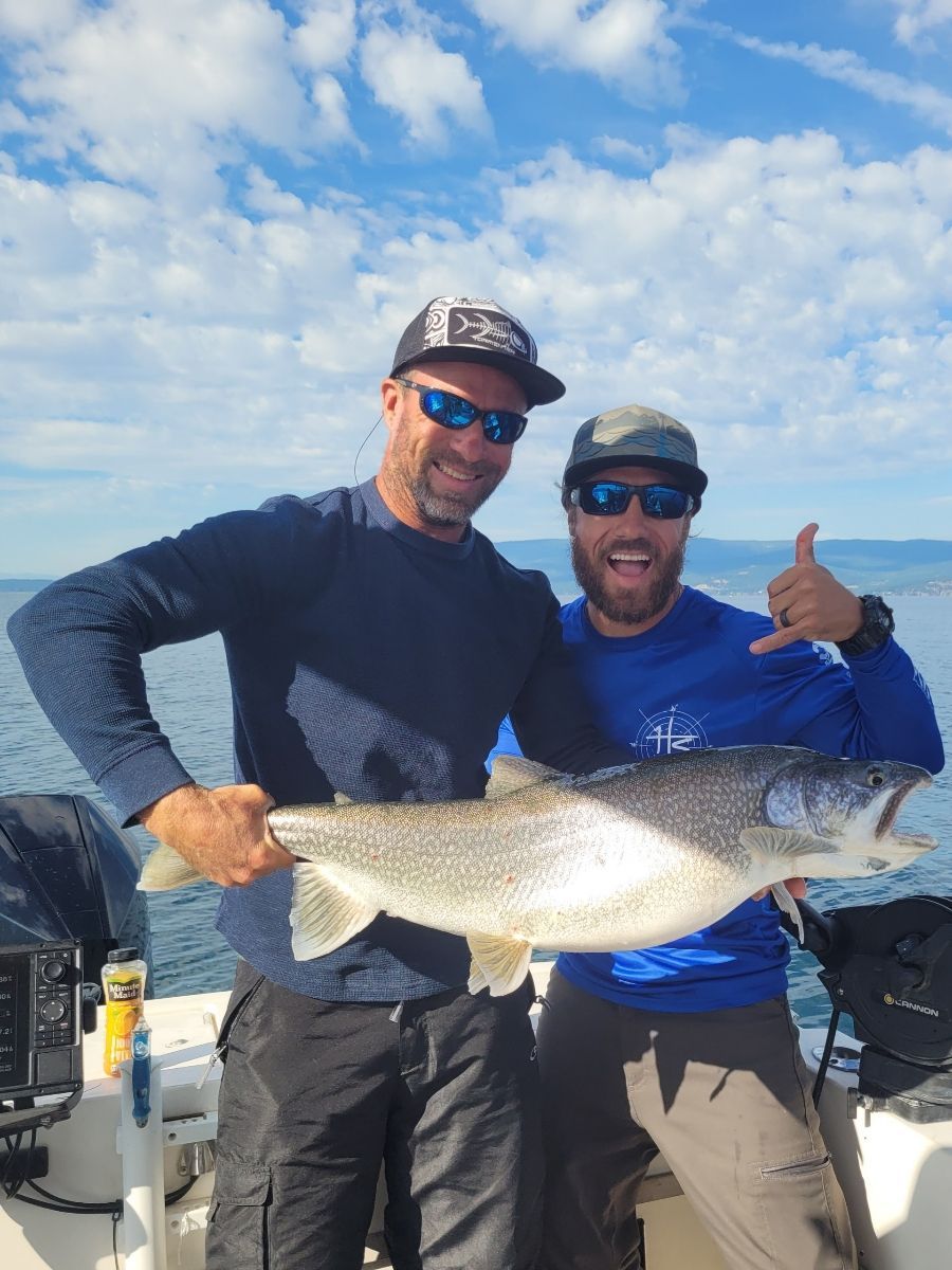 Two men on a boat, holding up a large fish they caught. Blue sky and water in background. One man gives a shaka.