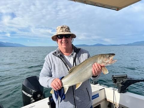 Man on boat holding a large fish, smiling. Lake and mountains in background.