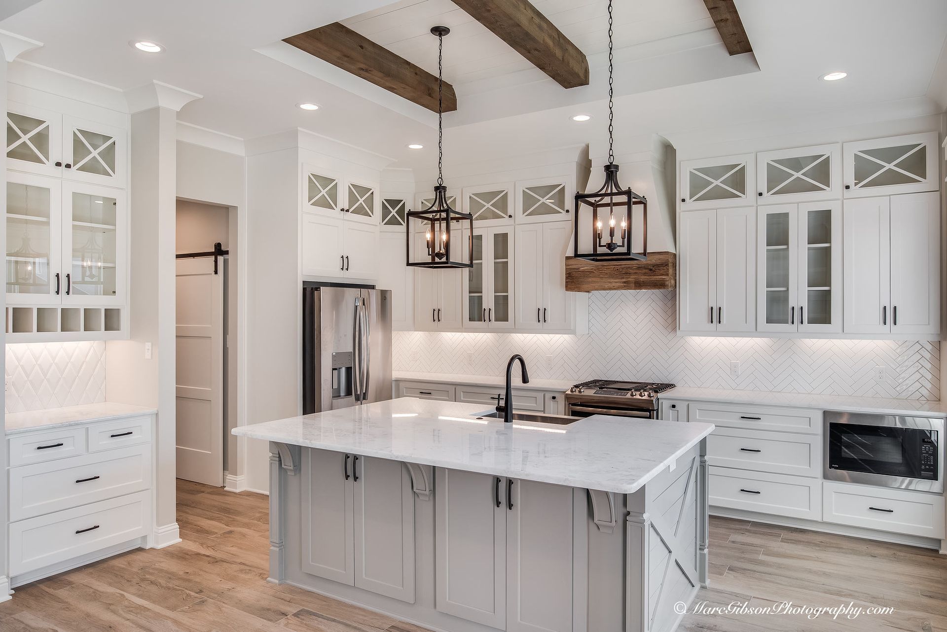 Modern kitchen with white cabinets, gray island, wooden beams, and stainless steel appliances.