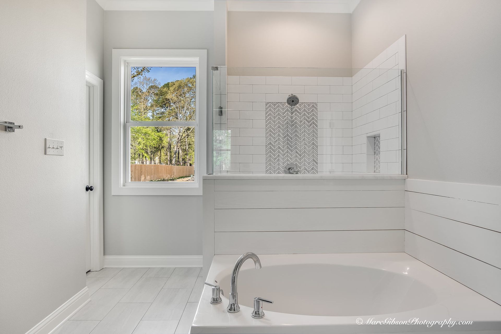 Bright bathroom with white tub, shower, and walls. A window offers an outside view.