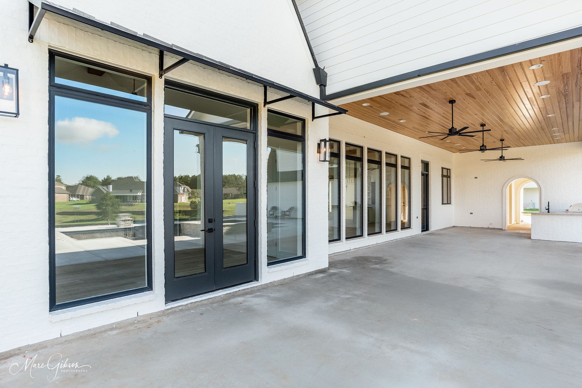 Exterior of a modern home with a concrete patio, black-framed windows, and a wood-paneled ceiling.