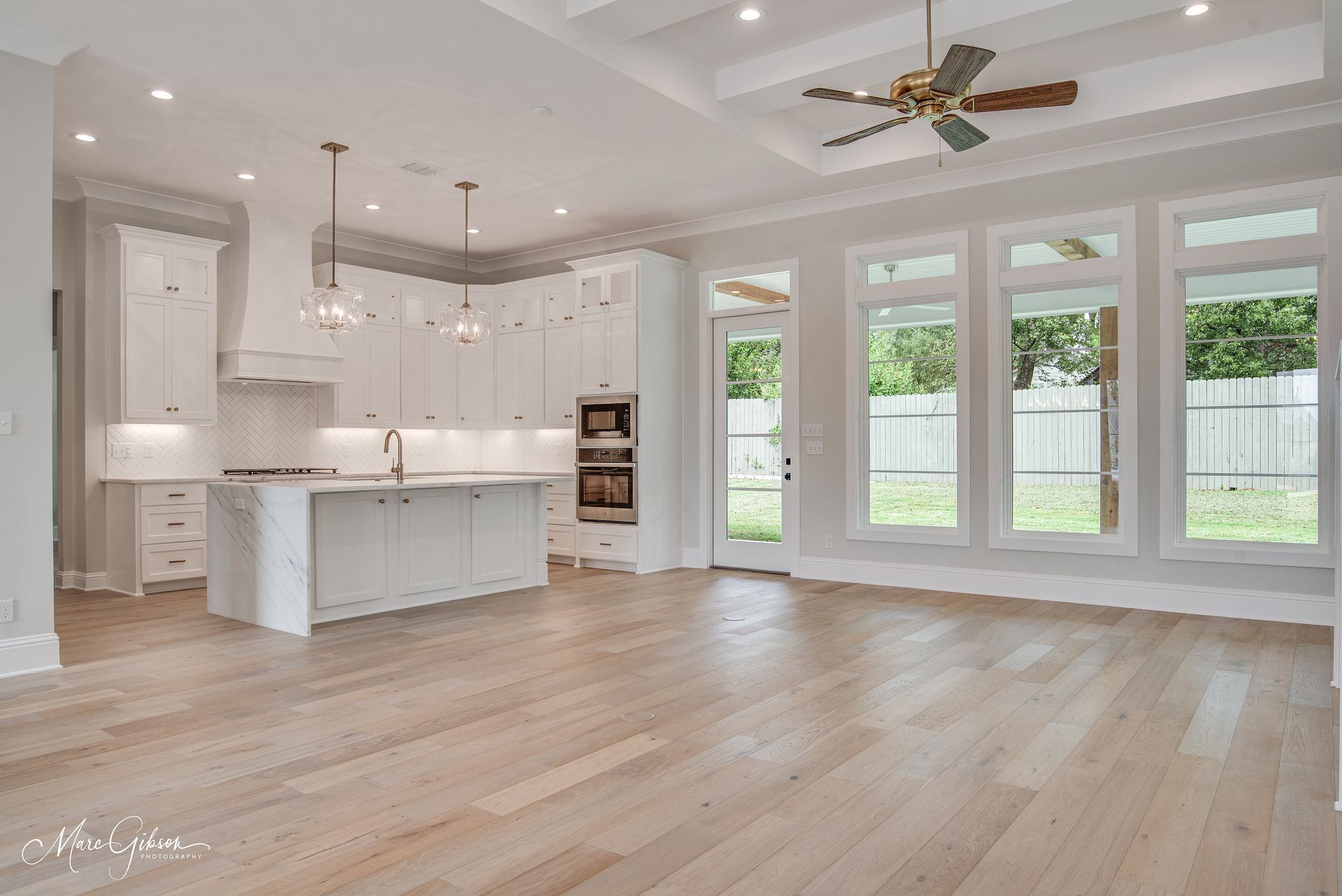 Spacious white kitchen with island, hardwood floors, and large windows overlooking a yard.