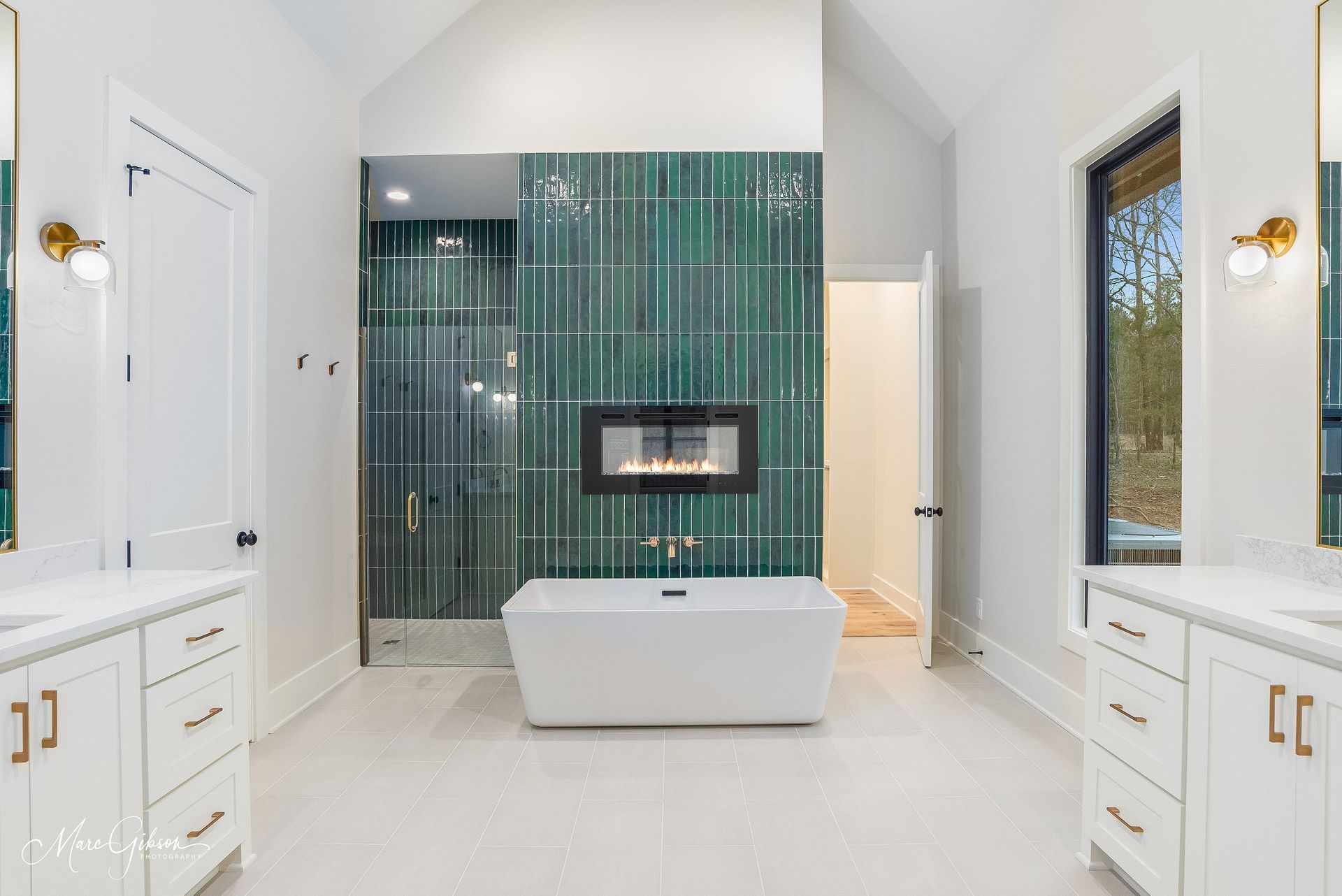 Modern bathroom with a green tiled wall, fireplace, white tub, and double vanity.