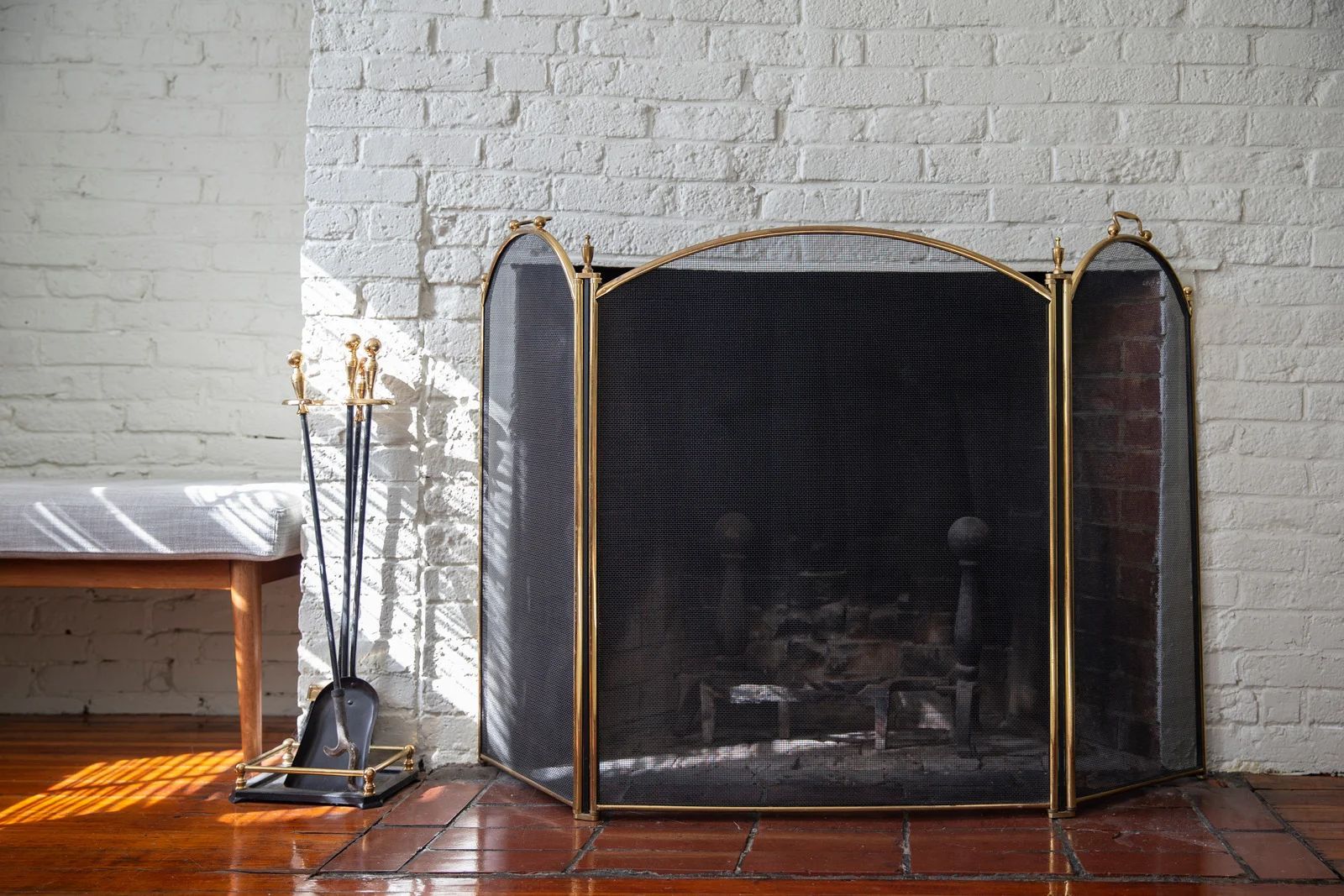 A three-paneled brass fireplace screen sits on a terracotta floor in front of a white brick wall and a small wooden bench.