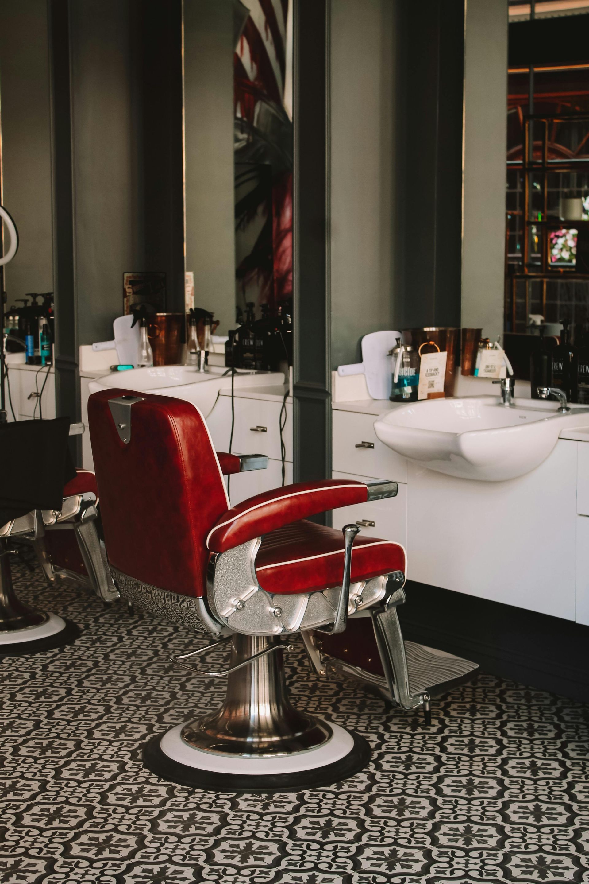Red barber chair in a vintage barbershop, surrounded by mirrors, sinks, and grooming products.