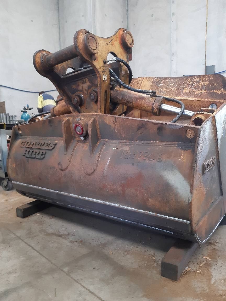 Rusty excavator bucket attachment in a workshop, showing hydraulic arms and worn metal surfaces.