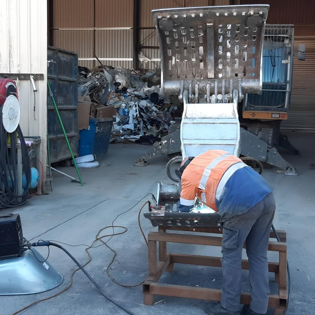 Person in a blue jacket bending over a workbench in a workshop, with metal sheets and equipment nearby.