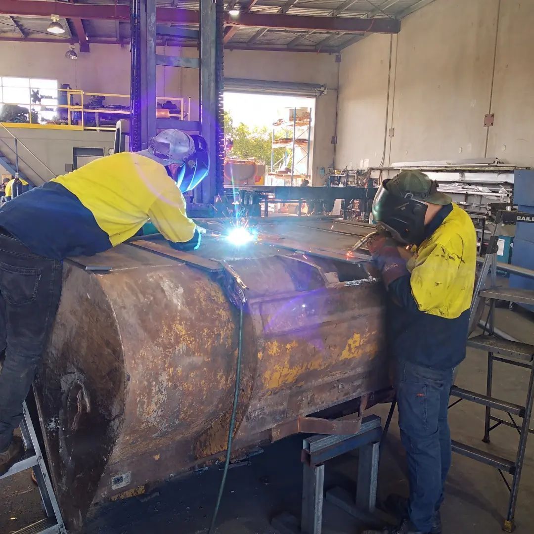 Two workers welding a large metal tank in a workshop, bright sparks visible.