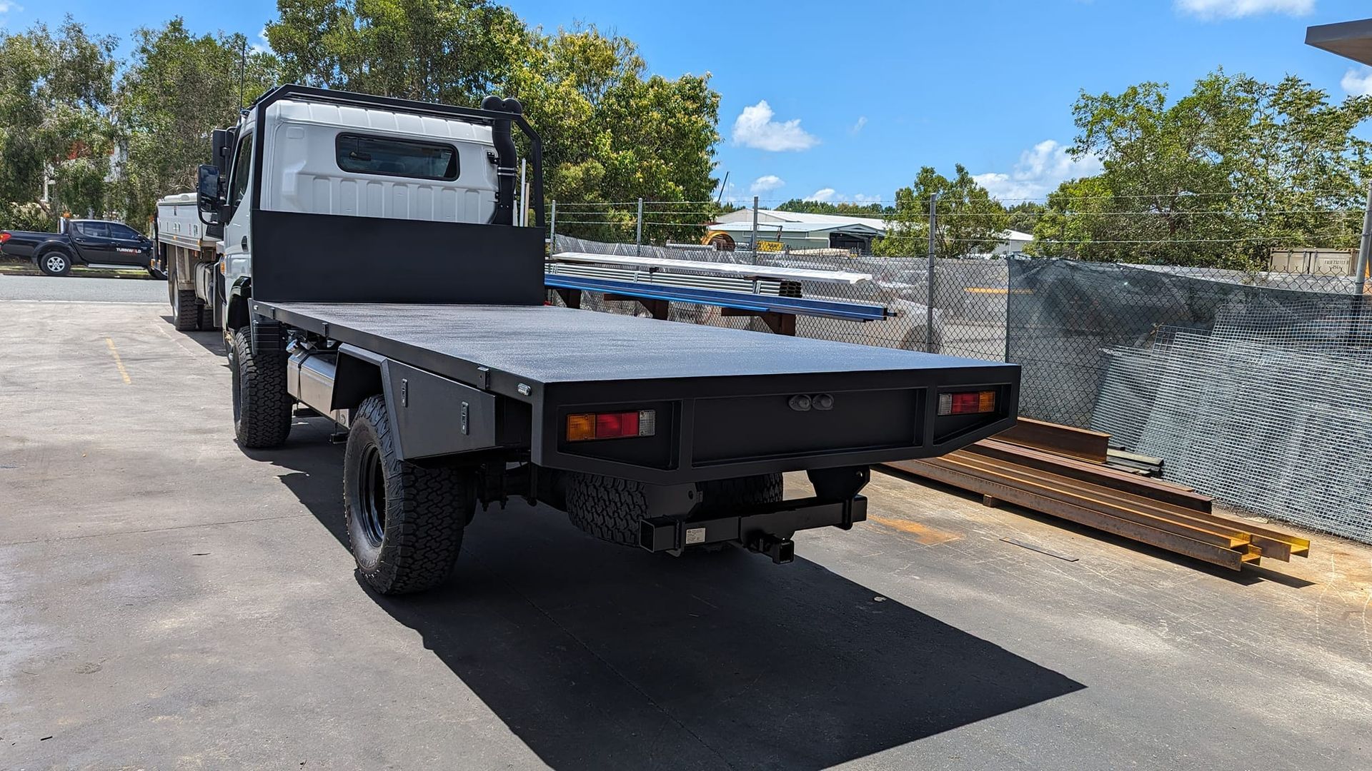 Flatbed work truck parked on a sunny street beside a chain-link fence and trees