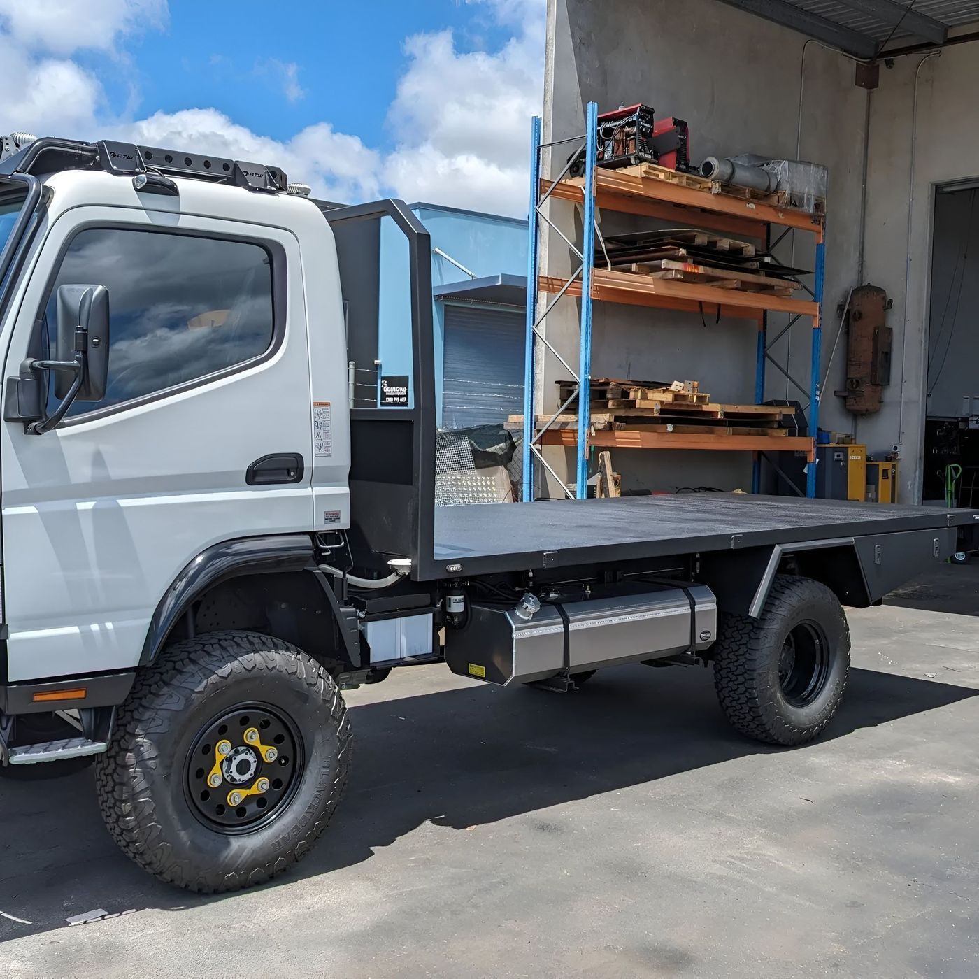 White flatbed truck parked outside a workshop with shelves of tools and equipment in the background