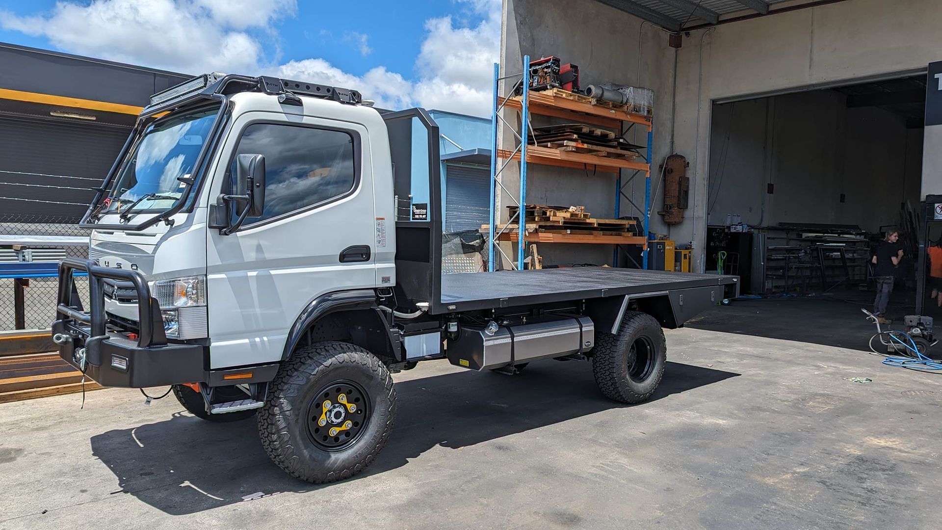 White flatbed truck parked beside a warehouse with shelving and a garage opening.