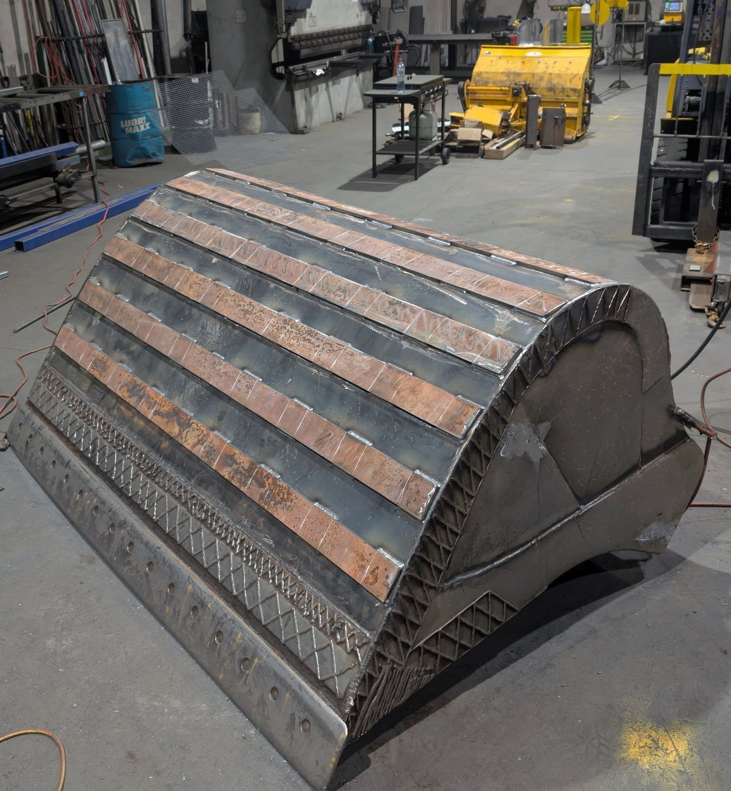 Large industrial metal bucket or crusher attachment in a workshop, with worn copper-colored wear strips on top.