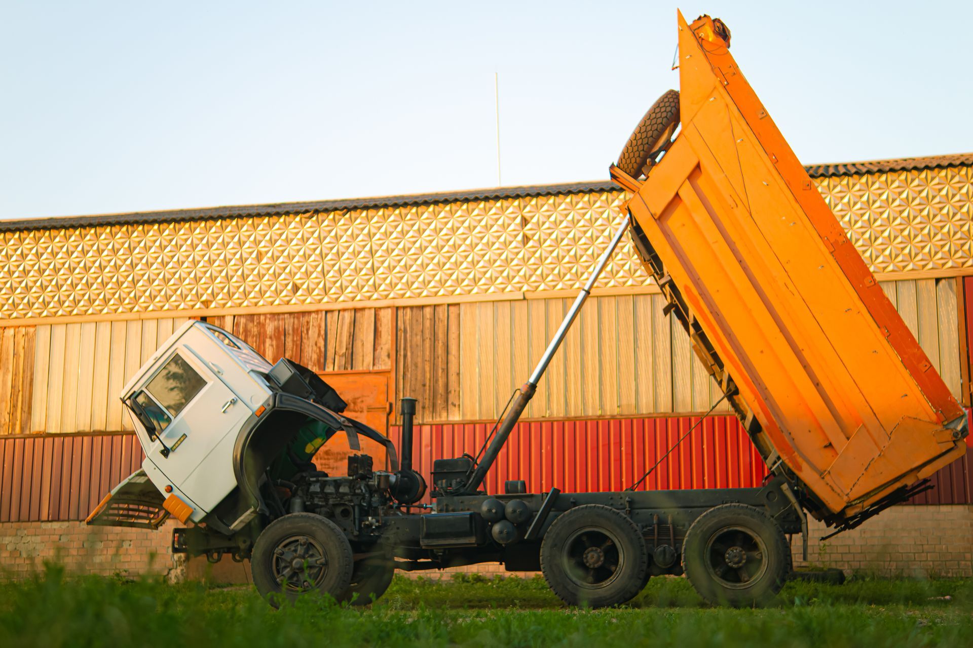 Overturned dump truck unloading orange trailer in a grassy field near a wooden fence