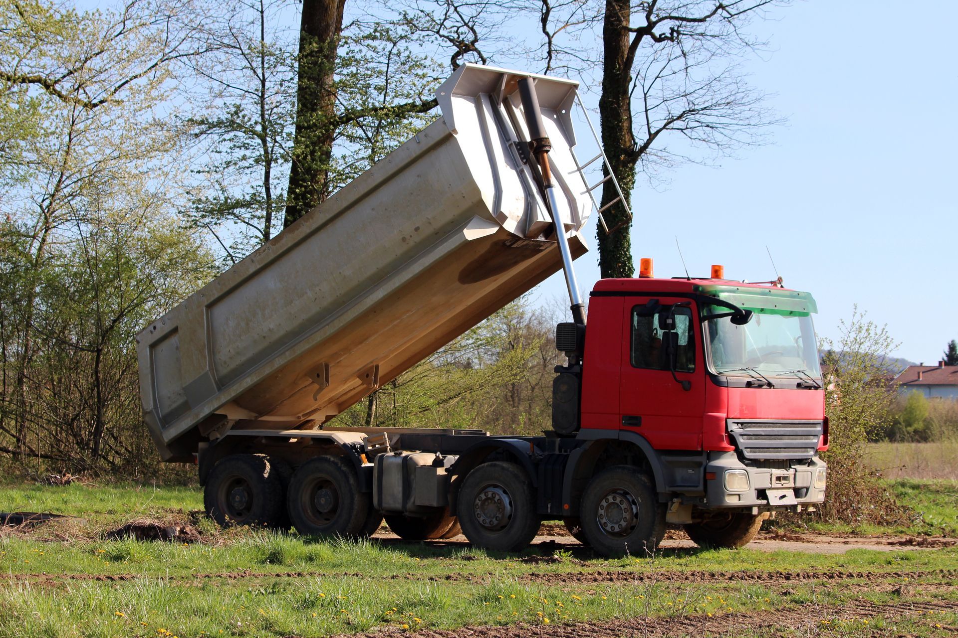 Red dump truck raises its bed to unload on a grassy roadside under trees.