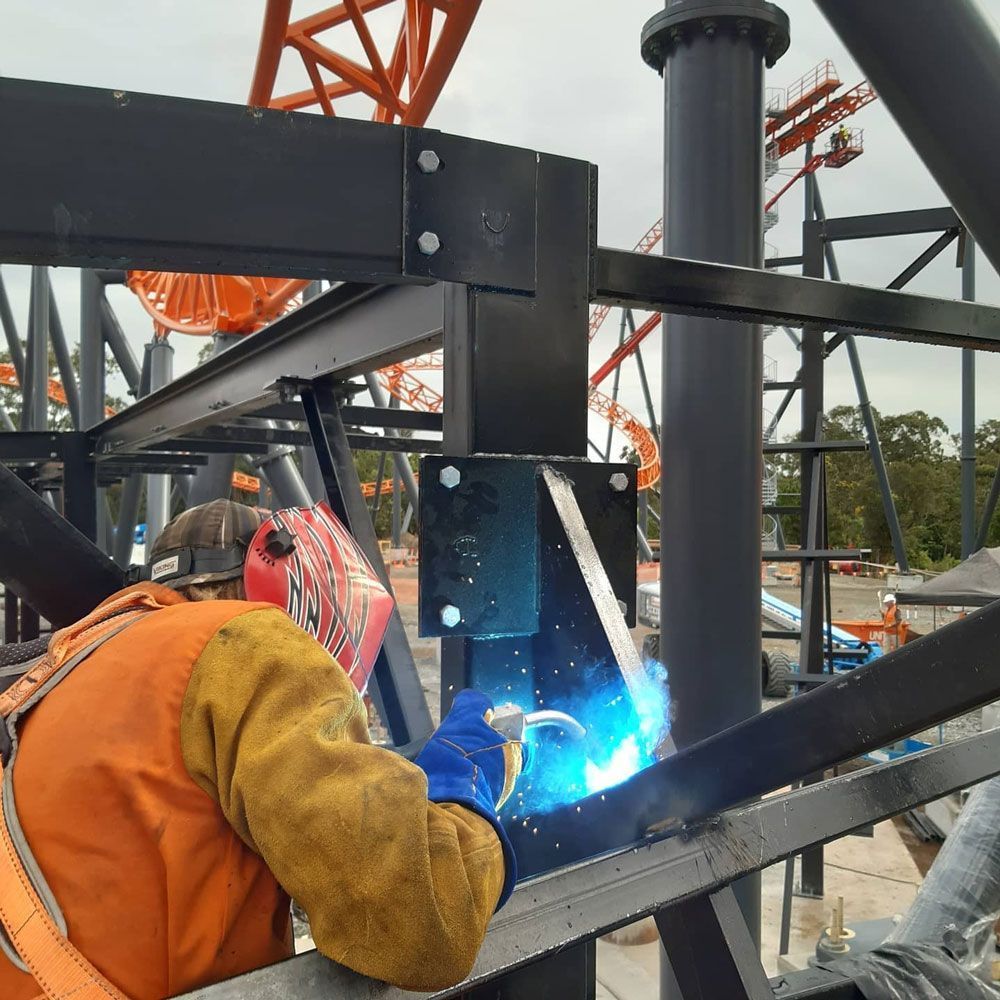 Worker welding a black steel frame, bright blue arc sparks in an industrial outdoor setting