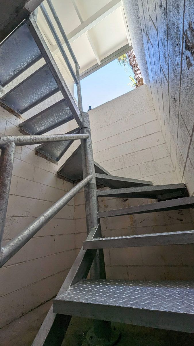 Metal staircase viewed from below, with blue handrails and sunlight from an opening above.