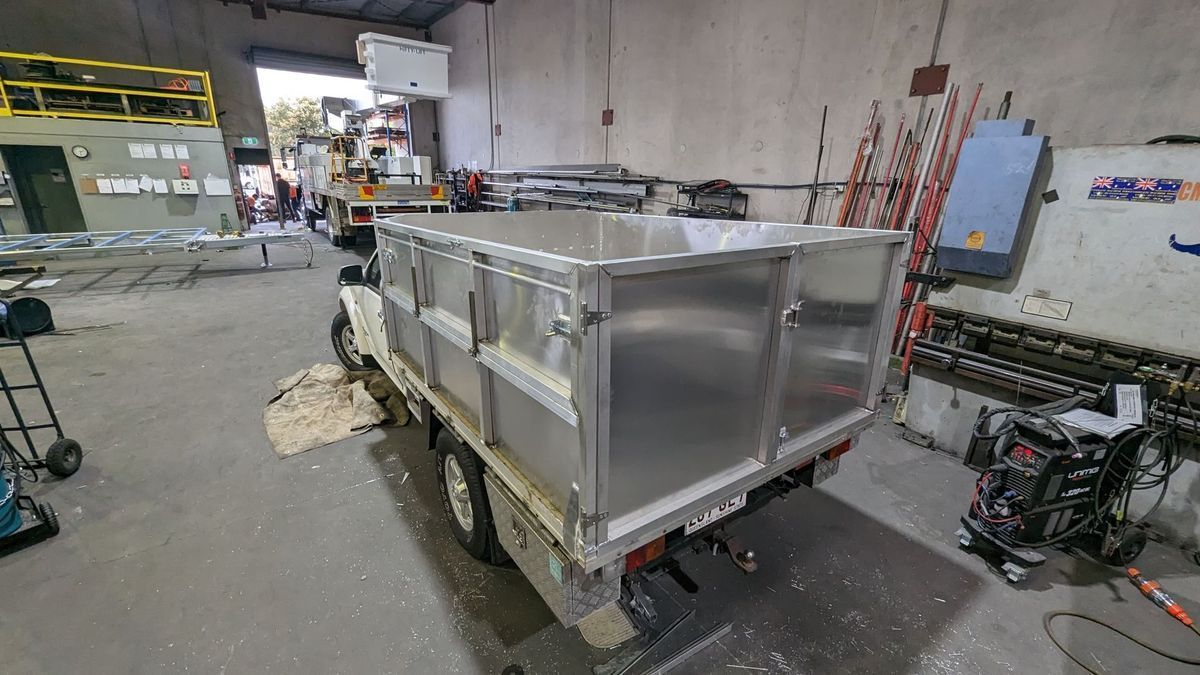 Metal utility trailer in a warehouse, with aluminum sides and a black hitch, surrounded by industrial equipment.