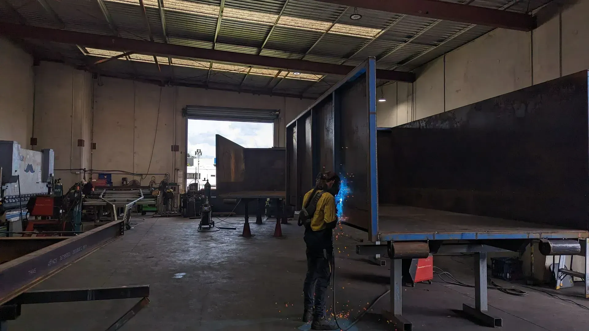 Person with yellow backpack in a warehouse beside a large black trailer and machinery.