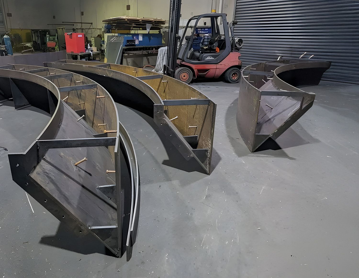 Workshop floor with several curved metal fabrication pieces and a forklift in the background